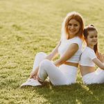 A mother and daughter sit back to back in matching activewear on a sunny grass field, enjoying a summer day together.