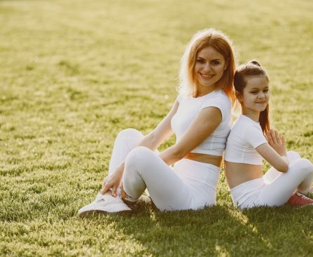 A mother and daughter sit back to back in matching activewear on a sunny grass field, enjoying a summer day together.