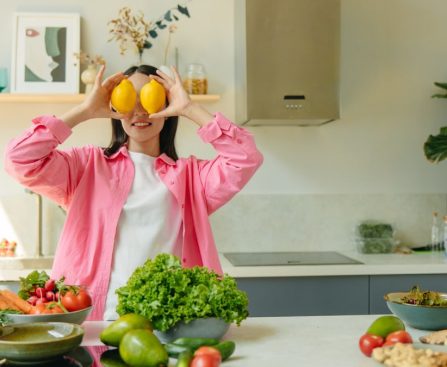 A woman playfully holding lemons over her eyes in a bright, lively kitchen setting filled with fresh vegetables.